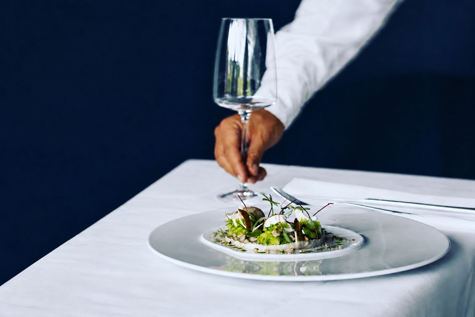 Ein Koch richtet ein Gourmetgericht auf einem weißen Tisch an, daneben steht ein Weinglas.