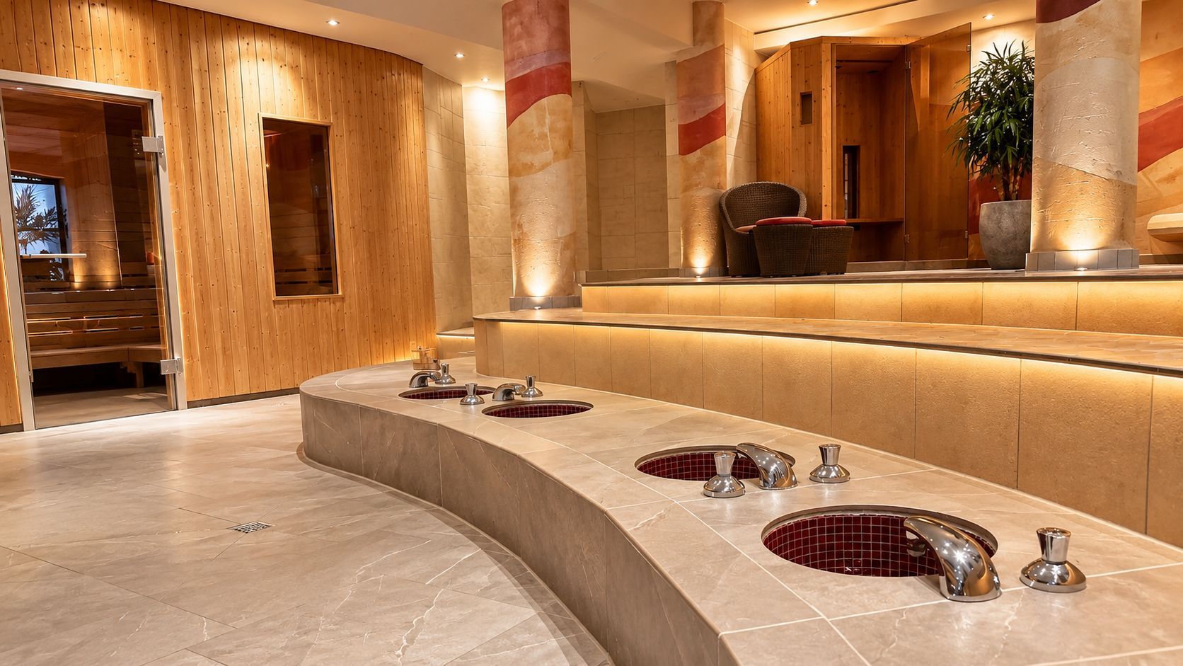 Luxuriöses Spa-Interieur mit geschwungenen Steinbänken, warmen Holzwänden und rot beleuchteten Fußbecken.