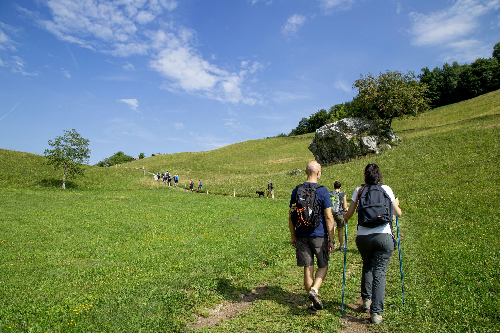 Zwei Wanderer durchqueren eine grüne Wiese unter blauem Himmel mit einem grasbewachsenen Hügel und vereinzelten Wolken.