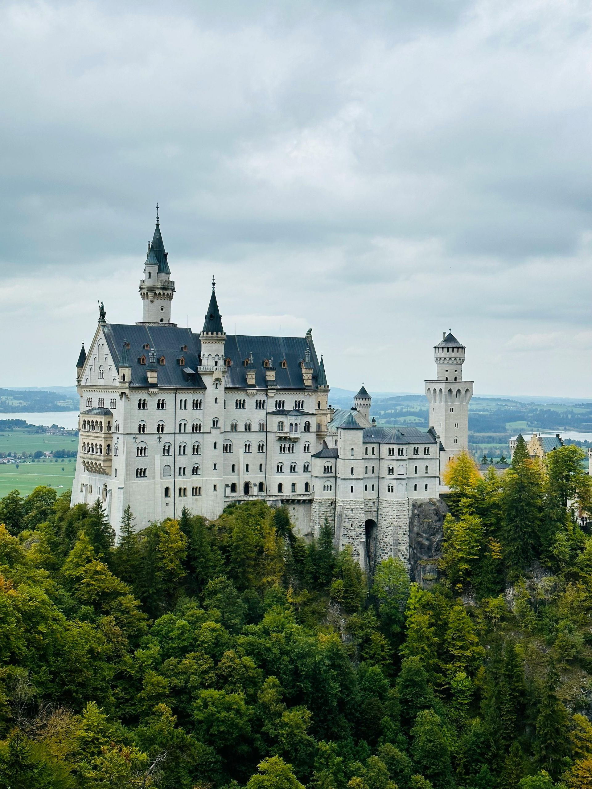 Weißes Schloss mit dunklen Dächern, das sich über grüne Bäume unter einem bewölkten Himmel erhebt