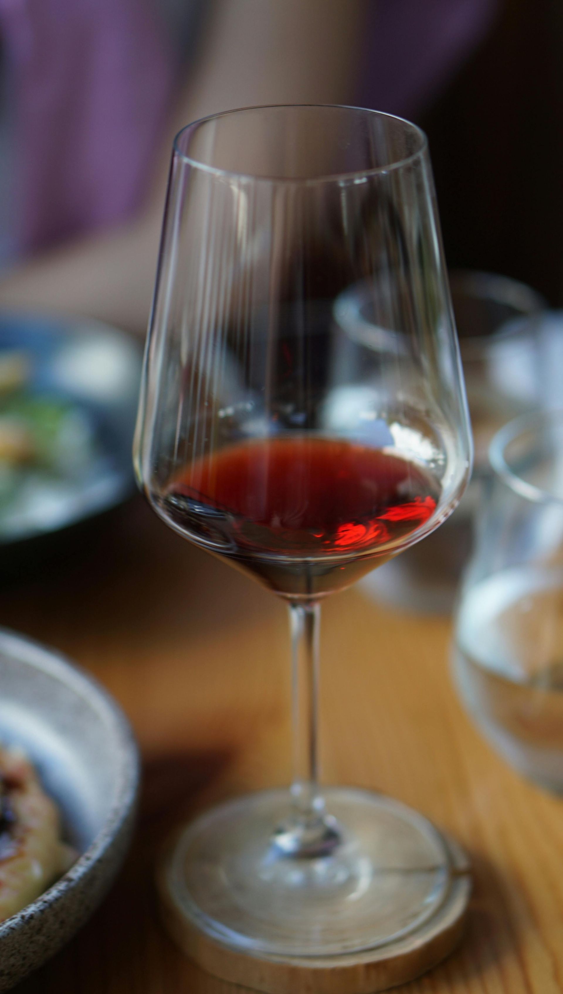 Ein Weinglas mit etwas Rotwein steht auf einem Tisch; im Hintergrund sind verschwommene Teller zu sehen.