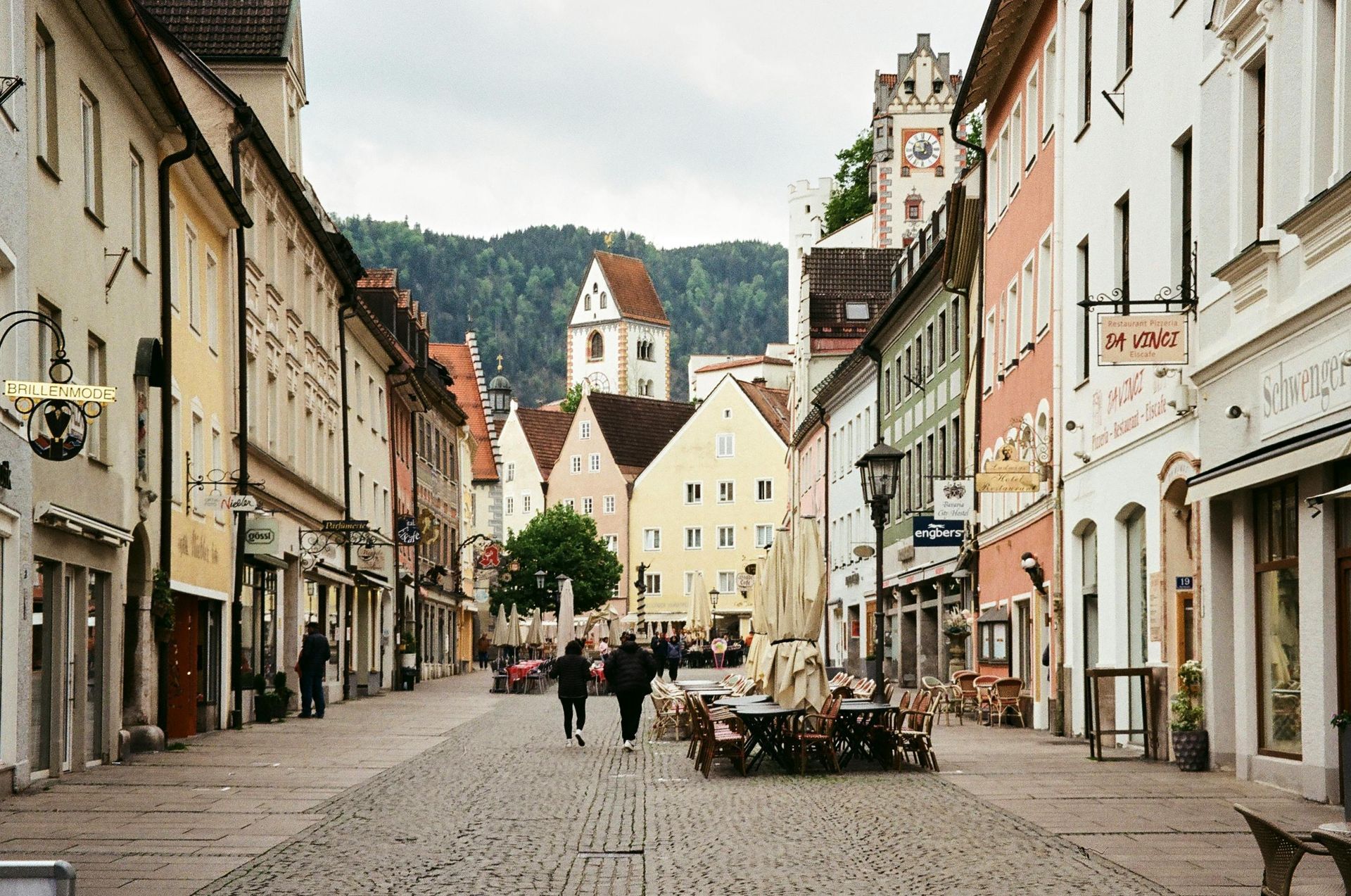 Kopfsteinpflasterstraße in einer europäischen Stadt mit historischen Gebäuden, Passanten und Hügeln im Hintergrund