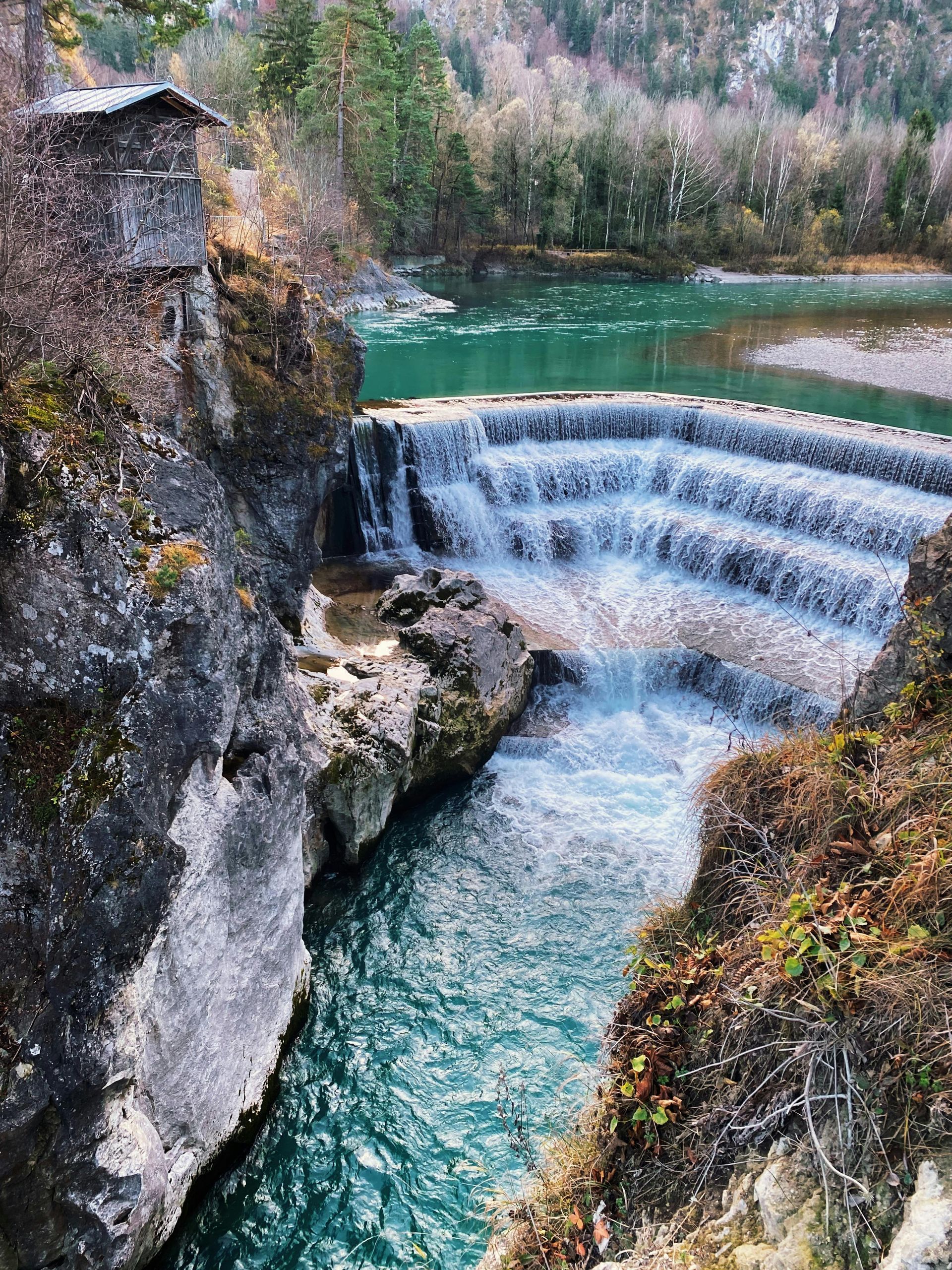 Ein terrassenförmiger Wasserfall stürzt zwischen Felswänden und immergrünem Wald in einen türkisfarbenen Fluss.