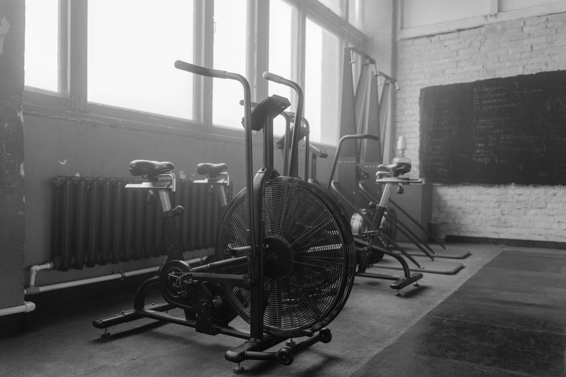 Ein Fitnessstudio in Graustufenoptik mit stationären Fahrradergometern, einem Airbike und großen Fenstern entlang der Wand.