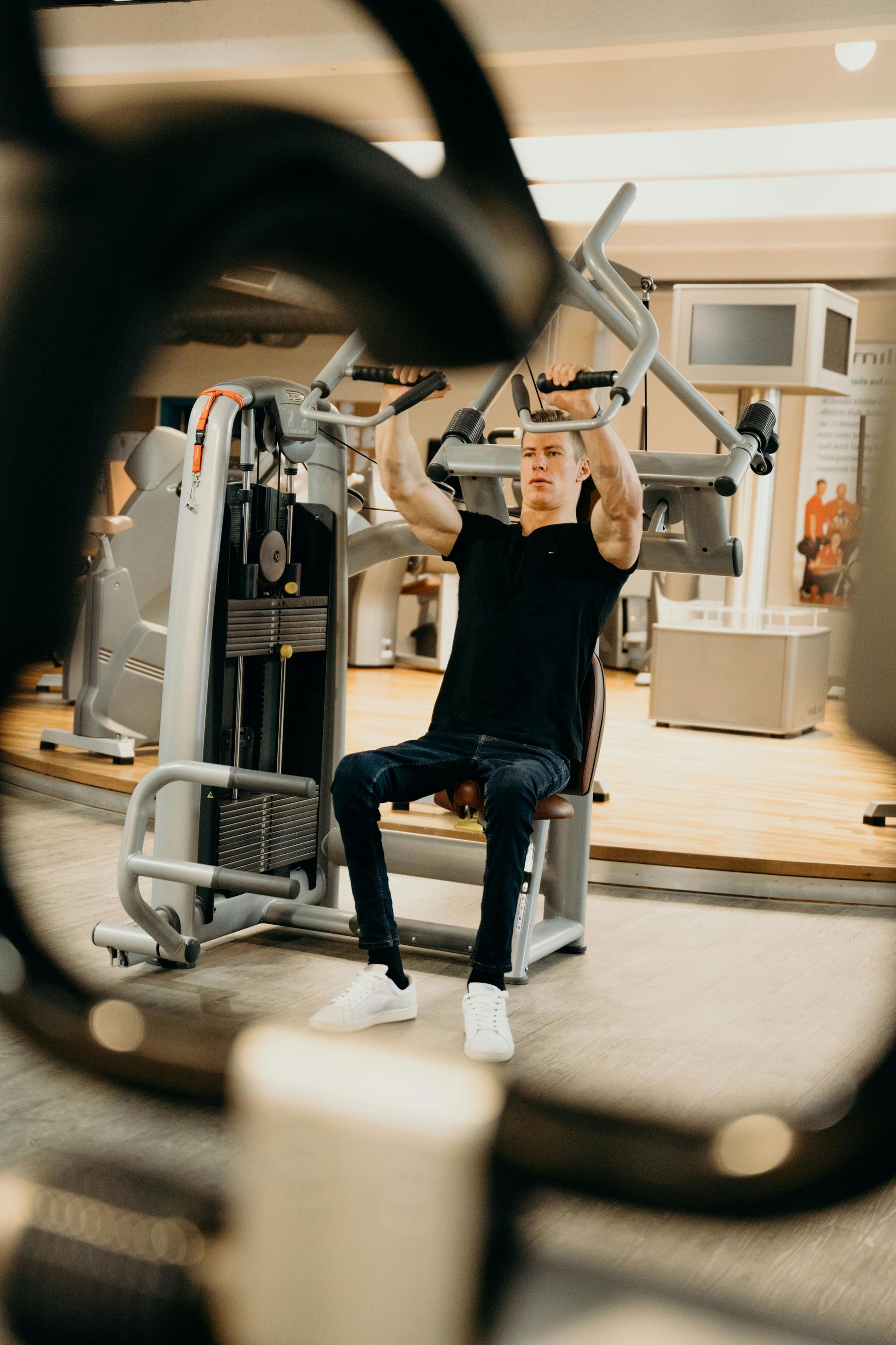 Eine Person benutzt ein Krafttrainingsgerät in einem Fitnessstudio; im Vordergrund sind verschwommene Geräte zu sehen.