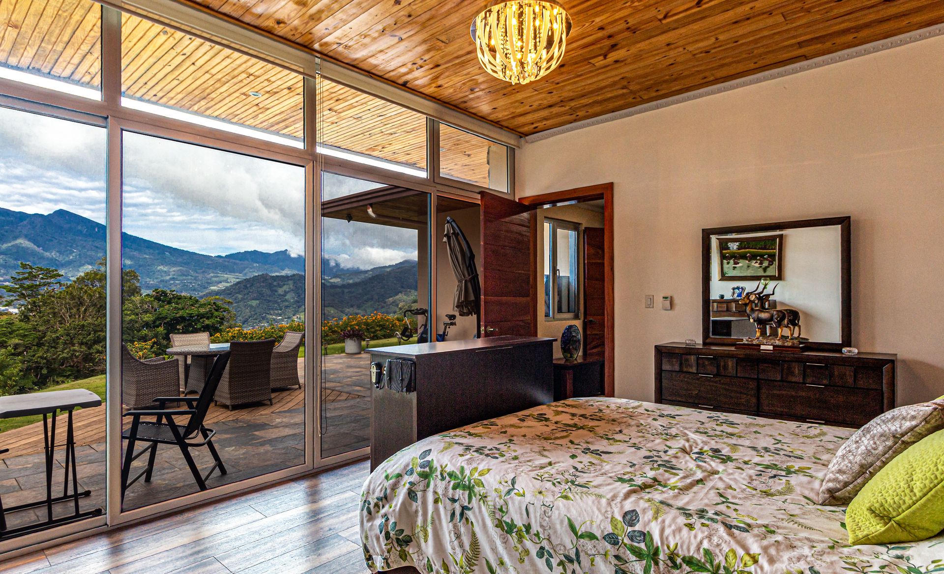Helles Schlafzimmer mit Balkonblick auf die Berge und den Sonnenuntergang, Holzdecke und geblümter Tagesdecke.