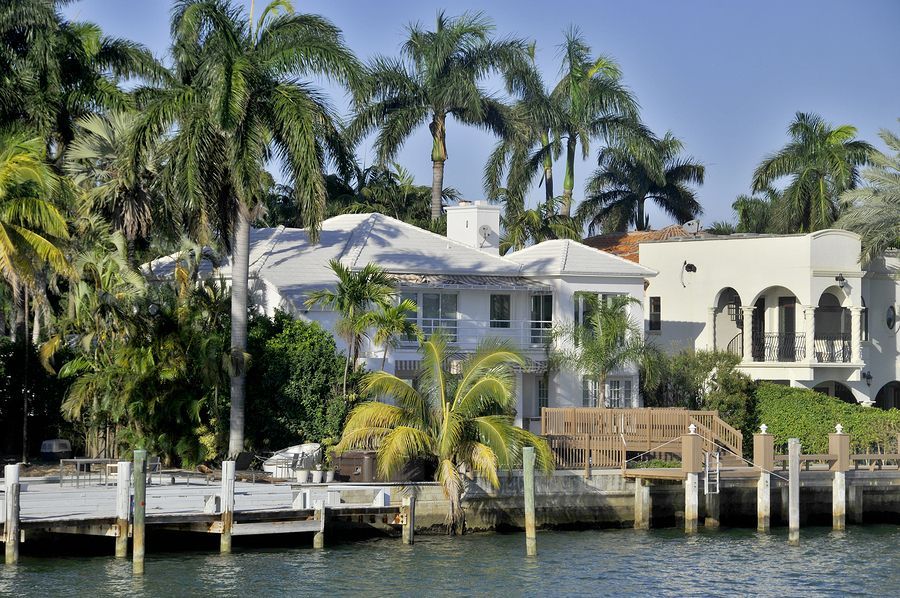 Waterfront white mansion with palm trees and a wooden dock.