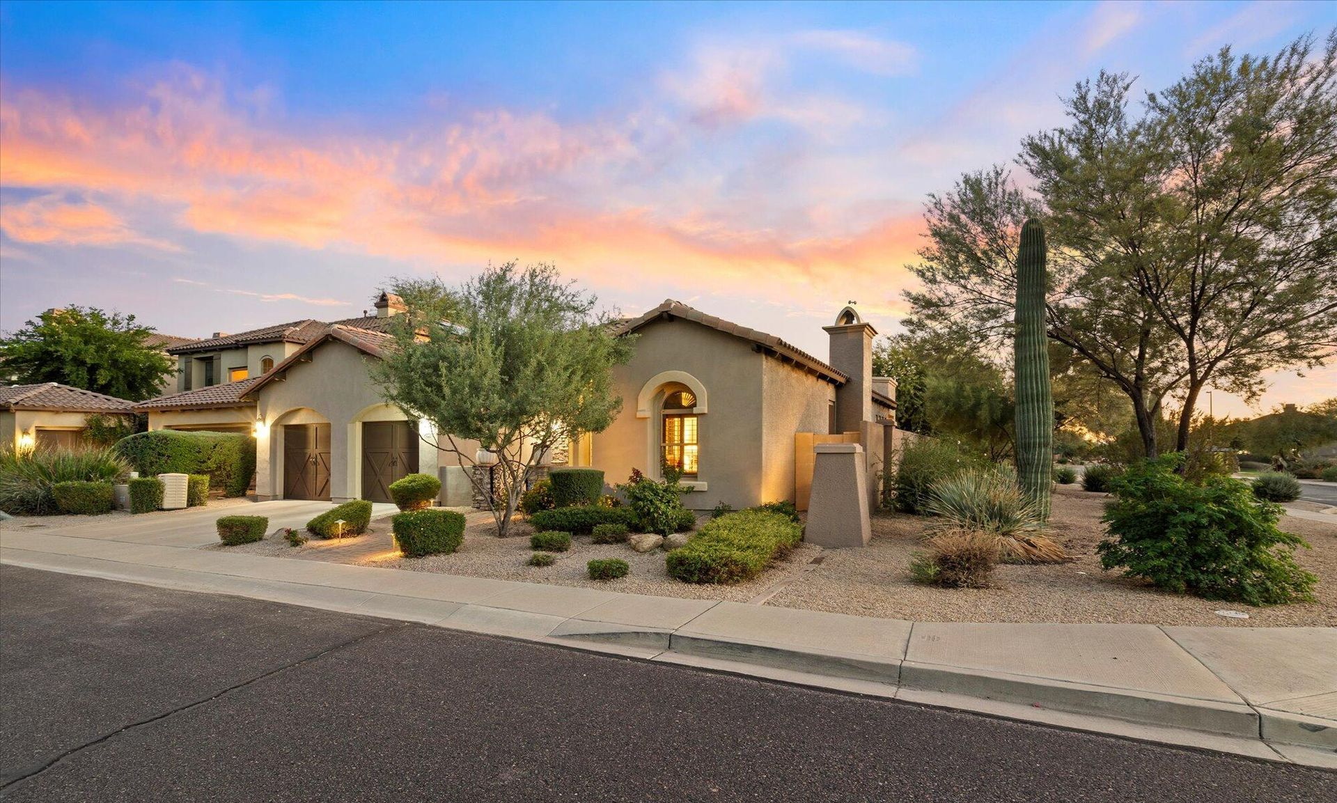 A beige-colored luxury home with lush desert landscaping under a colorful sunset sky.