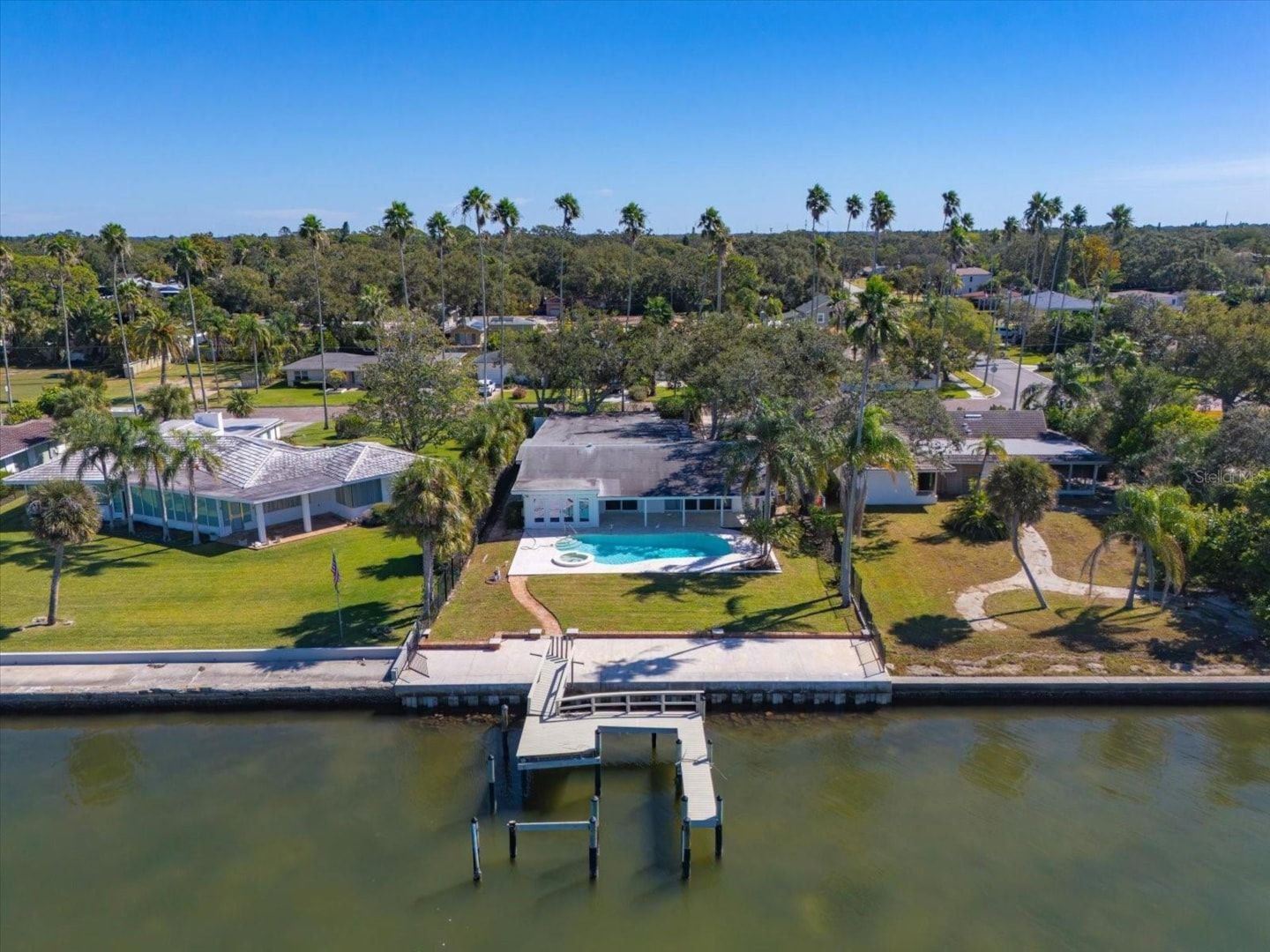 Waterfront home with pool and dock. Green grass, blue sky, palm trees.
