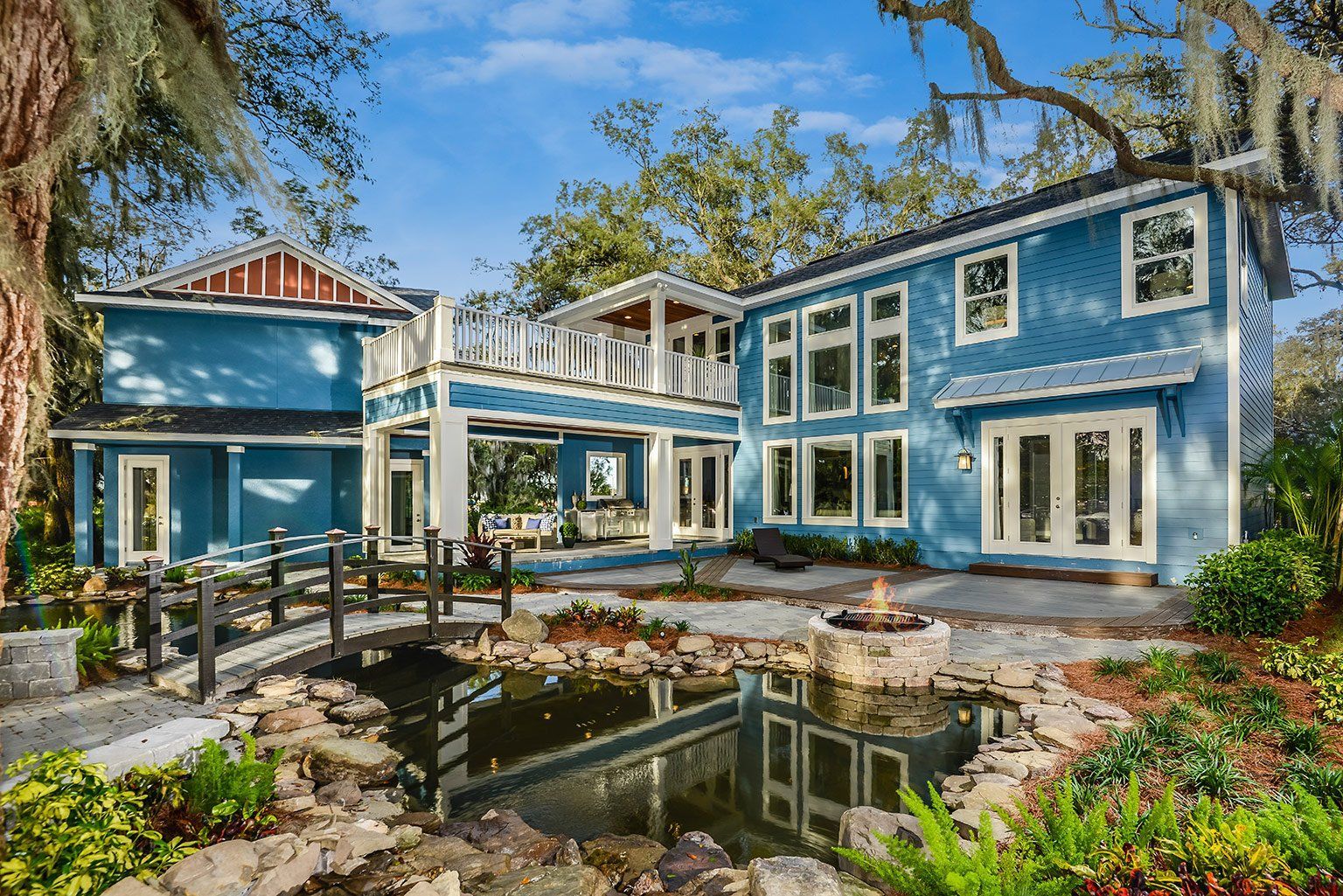 Blue two-story house with a white balcony and bridge over a small pond, surrounded by lush greenery under a blue sky.