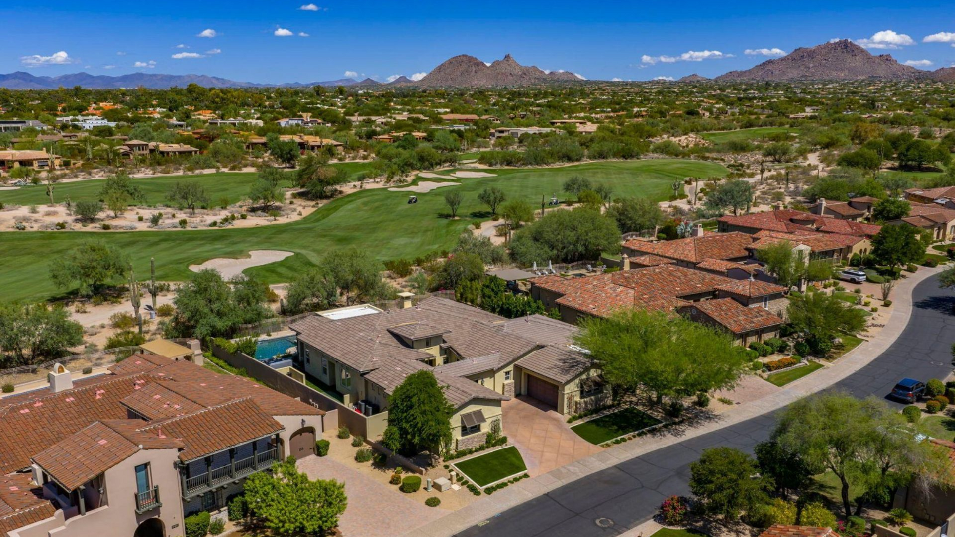 Aerial view of luxury homes with red-tile roofs near a golf course and mountains under a blue sky.