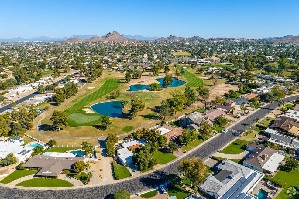 Aerial view of a golf course with ponds, surrounded by suburban homes and city landscape under a blue sky.