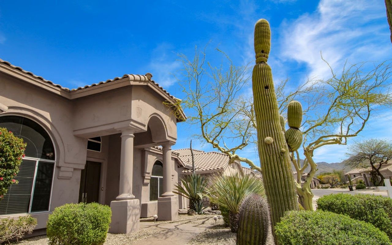 Beige house exterior with large saguaro cactus and desert plants under a blue sky.