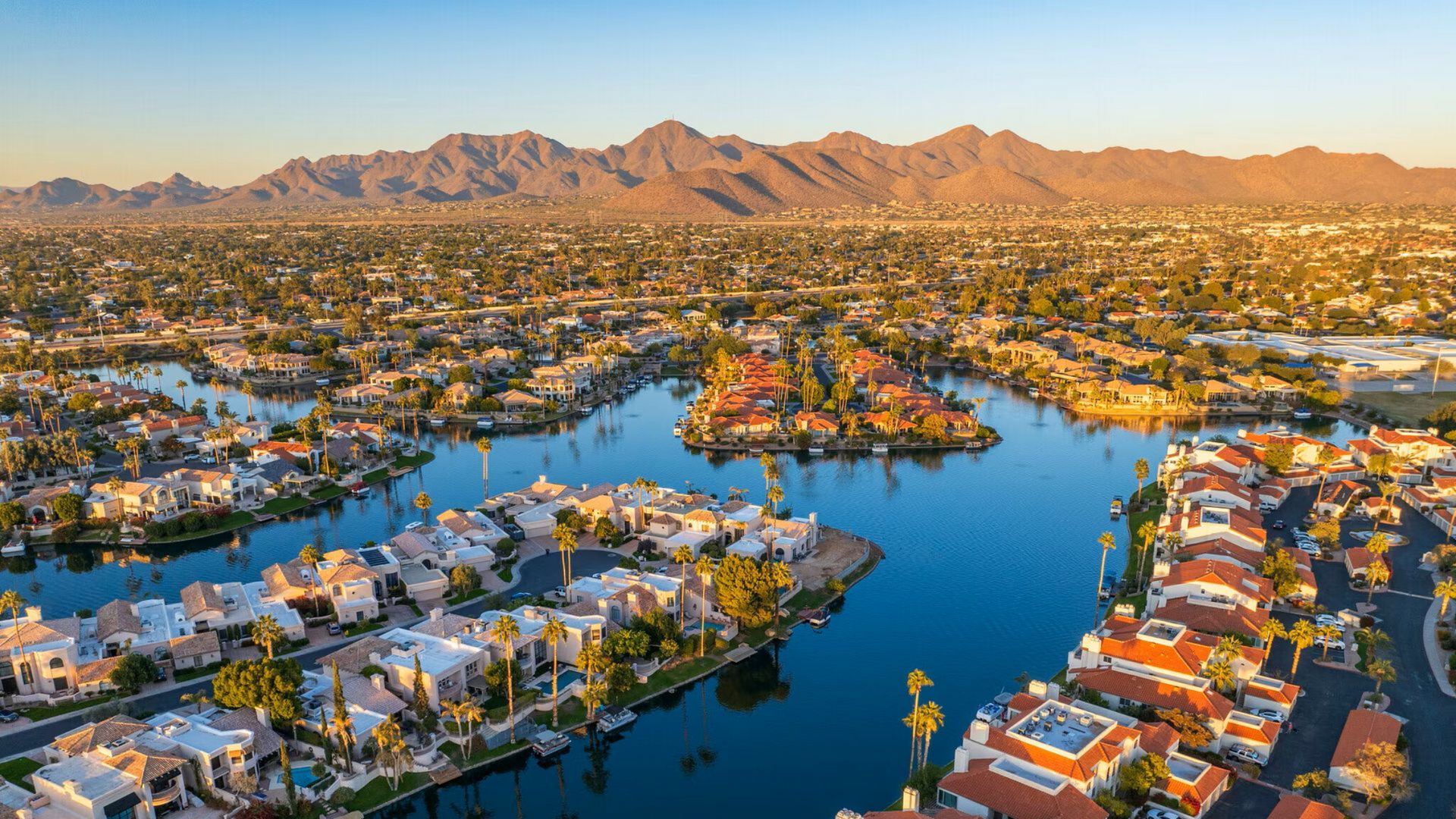 Aerial view of waterfront houses and canals in front of a mountain range, bathed in golden sunlight.