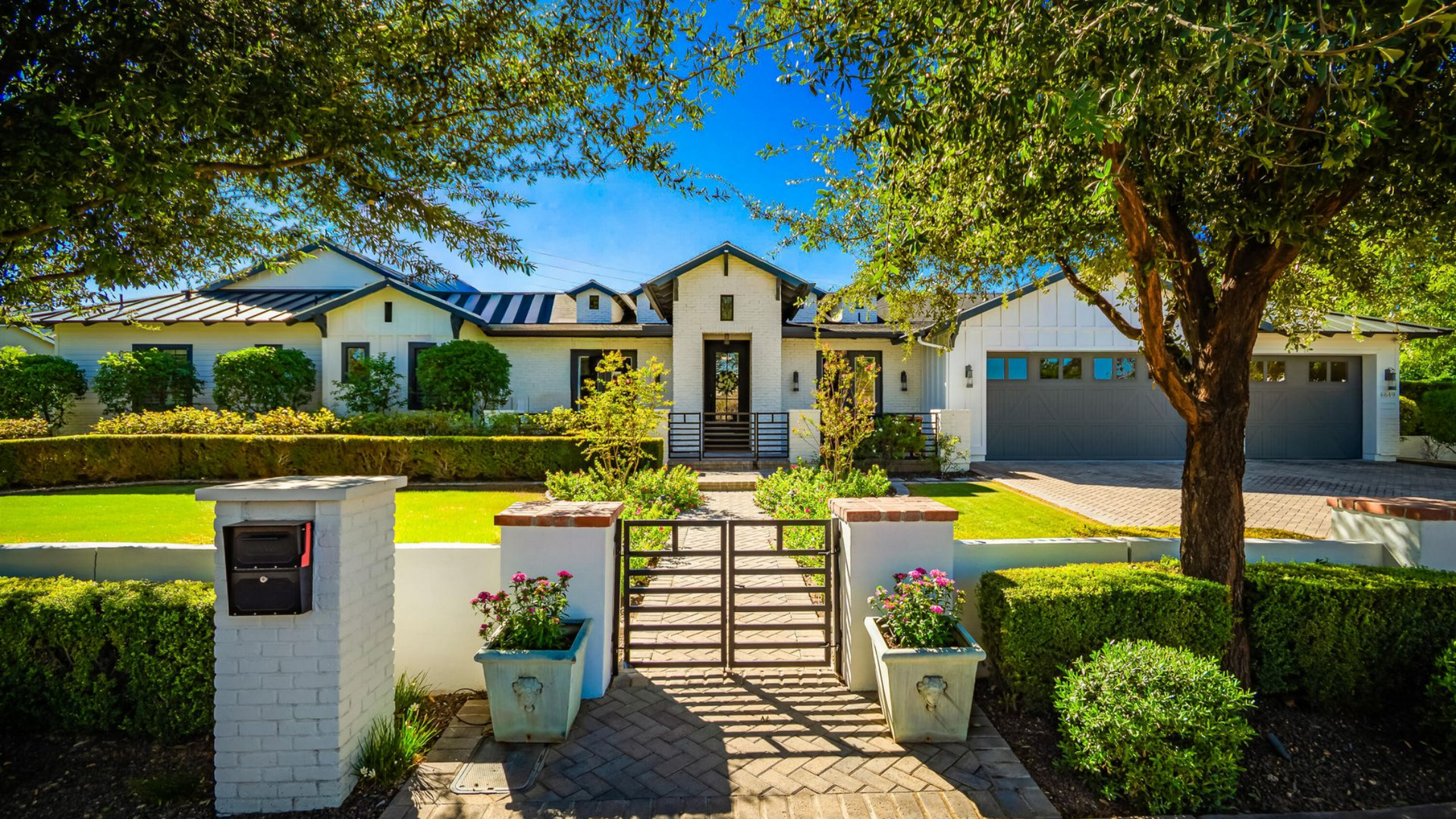 White house with black accents, lush green lawn, trees, gate, and a blue sky.