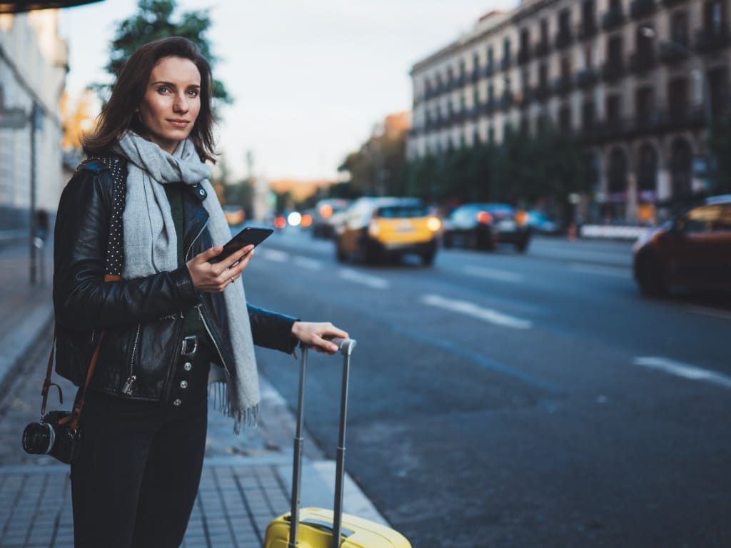 Una mujer está parada al costado de la carretera con una maleta y mirando su teléfono.