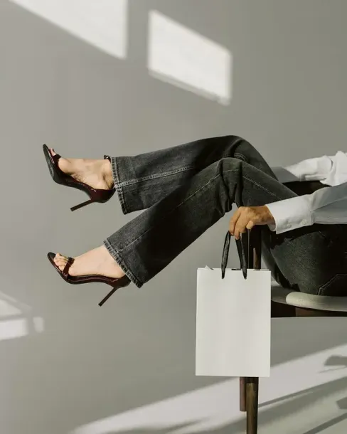 Una mujer está sentada en una silla con las piernas cruzadas y sosteniendo una bolsa de compras.