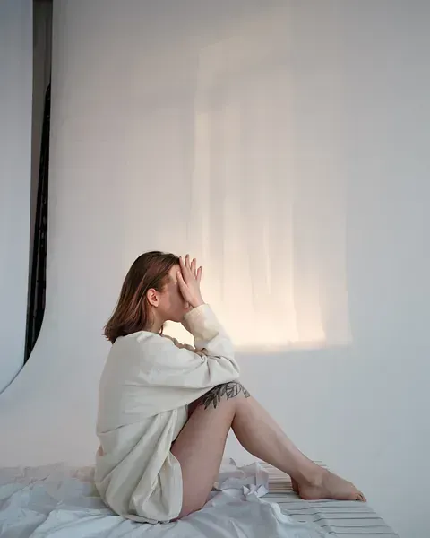 Una mujer está sentada en una cama cubriéndose la cara con las manos.
