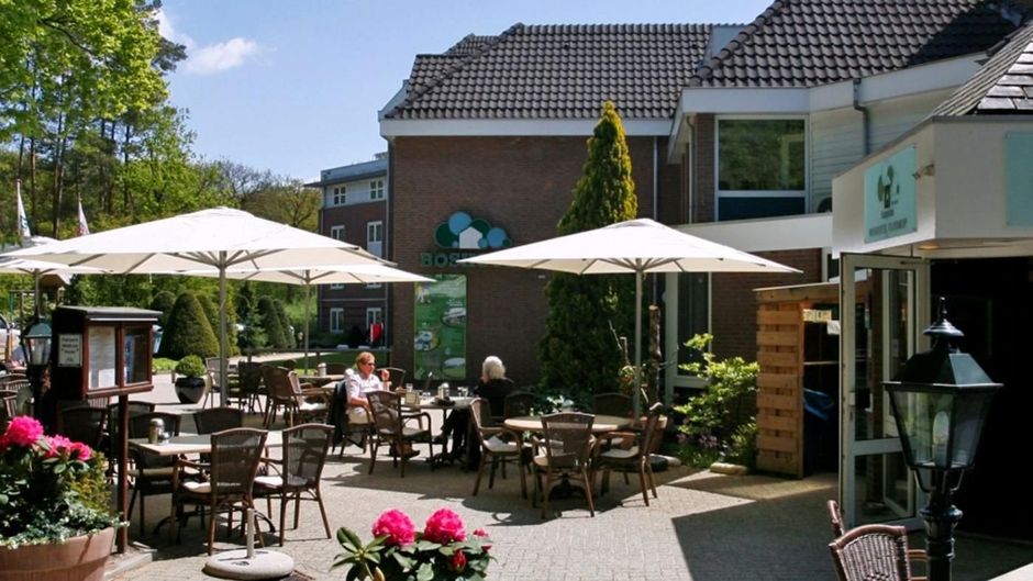Terras van de Brasserie met tafels, witte parasols en bloemen naast een gebouw op een zonnige dag.
