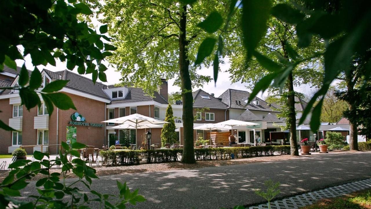 A restaurant with tables and umbrellas in front of a building surrounded by trees.