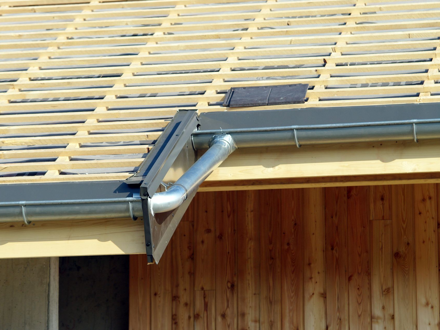 Toiture inachevée avec chevrons en bois, gouttière et descente pluviale argentée.