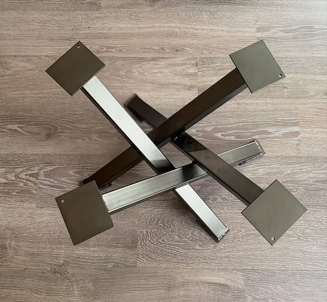 Quatre pieds de table en métal avec plaques de montage carrées, croisés sur un plancher en bois.