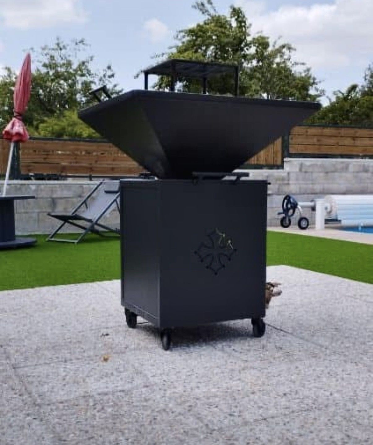 Barbecue d'extérieur en métal noir avec roulettes, façade décorative et base carrée, posé sur une terrasse avec de l'herbe verte.