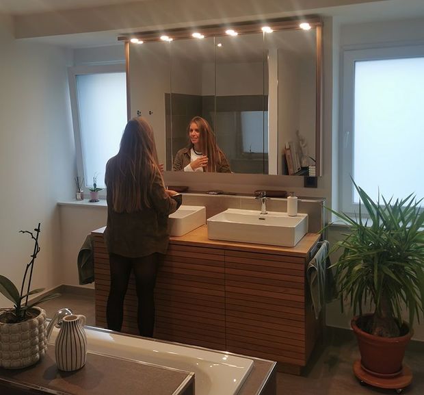 Eine Person steht an einem Waschtisch im Badezimmer mit Spiegel, Waschbecken und Topfpflanzen in einem sanft beleuchteten Raum.