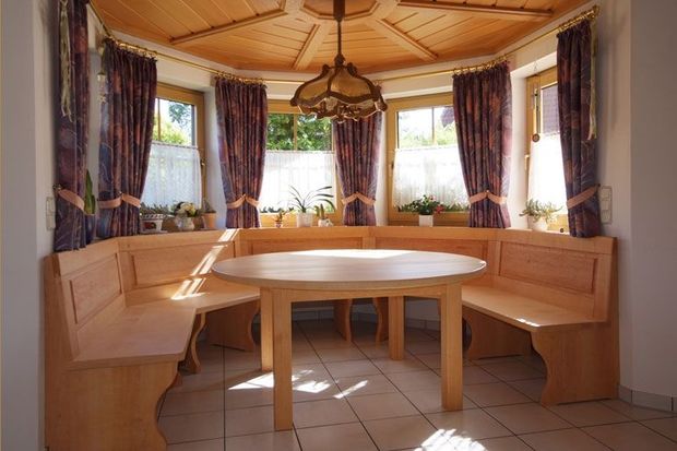 Gemütliche Frühstücksecke aus Holz mit eingebauten Bänken, rundem Tisch und Vorhängen um helle Fenster.