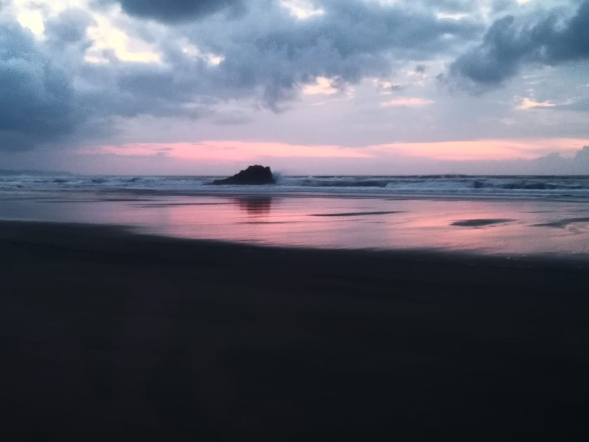 Playa al anochecer, cielo rosa y morado reflejado en la arena mojada, nubes oscuras arriba, pequeña roca en el océano.