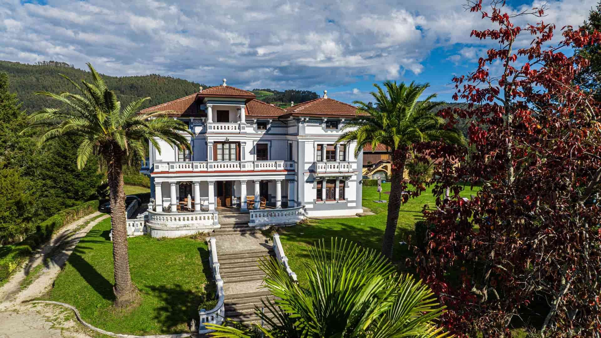 Elegante mansión blanca con detalles azules, palmeras y césped verde, ubicada en un paisaje montañoso.