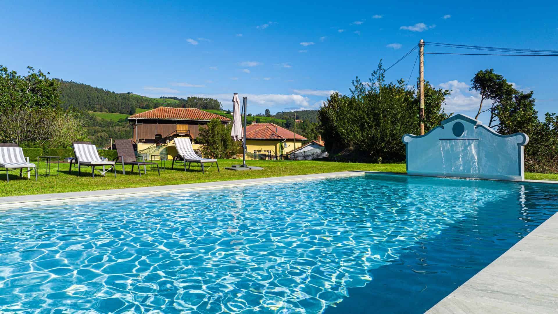 Piscina con césped circundante y tumbonas, con casas a lo lejos y cielo azul.