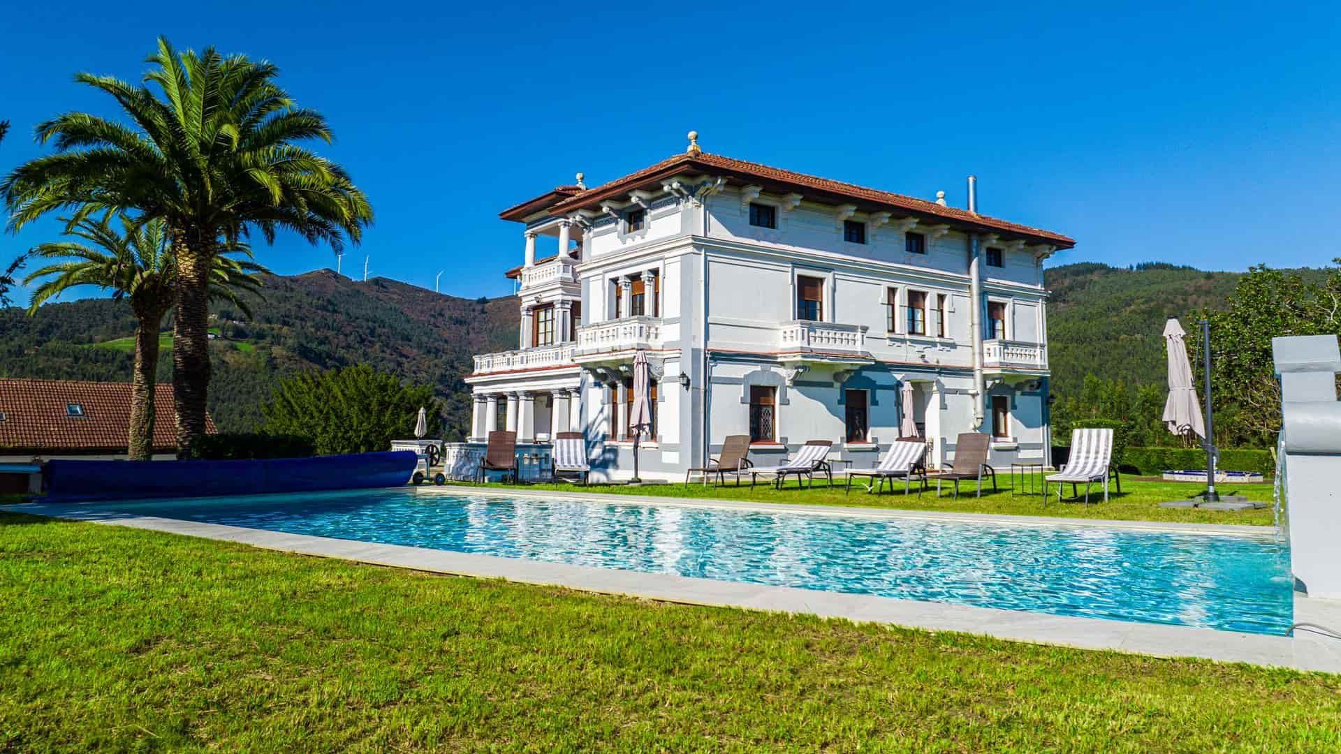 Villa blanca con piscina, palmeras y fondo de montaña en un día soleado.