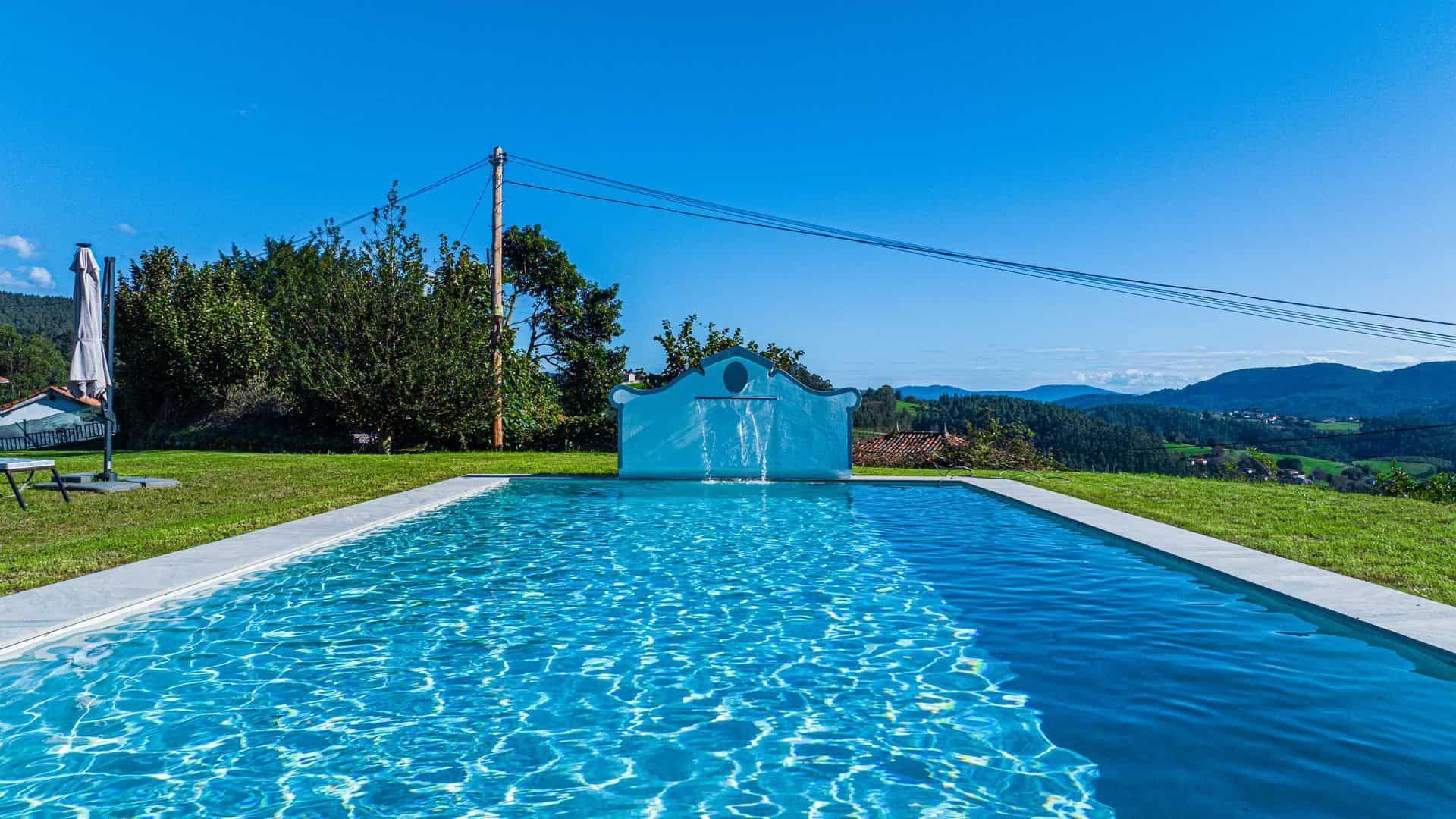 Piscina con agua azul, bordes blancos y una pequeña estructura blanca en una zona de césped con vista a la montaña.