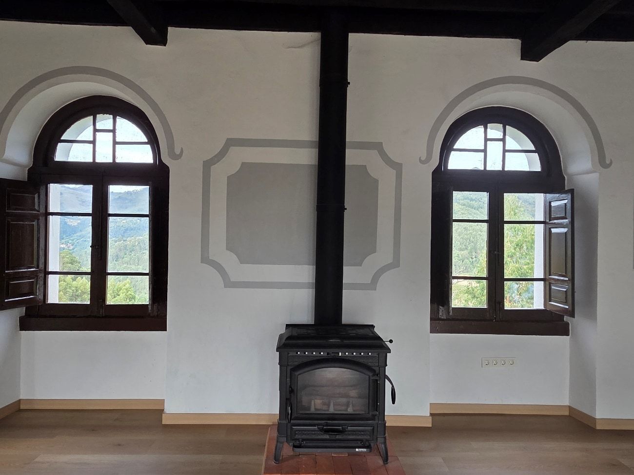 Habitación con ventanas arqueadas, chimenea y chimenea contra una pared blanca; contraventanas de madera abiertas, con vista a la vegetación.