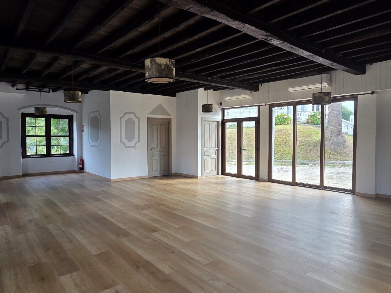 Habitación vacía con suelo de madera, vigas oscuras en el techo, ventanas, puertas y paredes blancas.