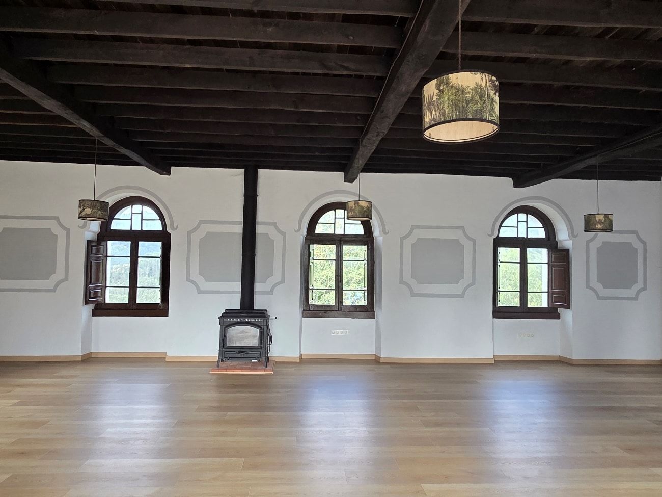 Habitación espaciosa con suelo de madera, ventanas arqueadas y vigas de madera oscura. Una estufa de leña se encuentra en el centro.