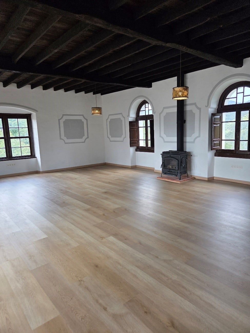 Habitación vacía con suelo de madera, ventanas arqueadas con persianas y vigas de madera oscura en el techo. Chimenea y lámparas.