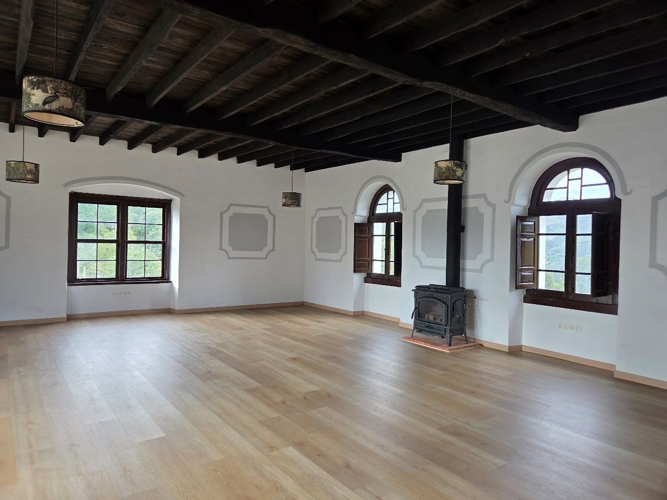 Habitación vacía con piso de madera, paredes blancas, techo de madera oscura, ventanas arqueadas y chimenea.