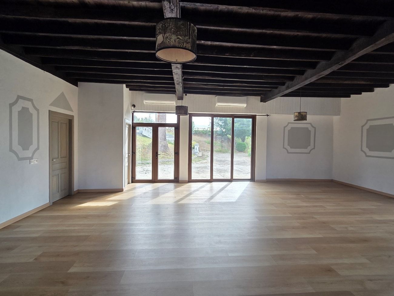 Habitación vacía con piso de madera, ventanas grandes, vigas oscuras en el techo y marcos de pared decorativos.