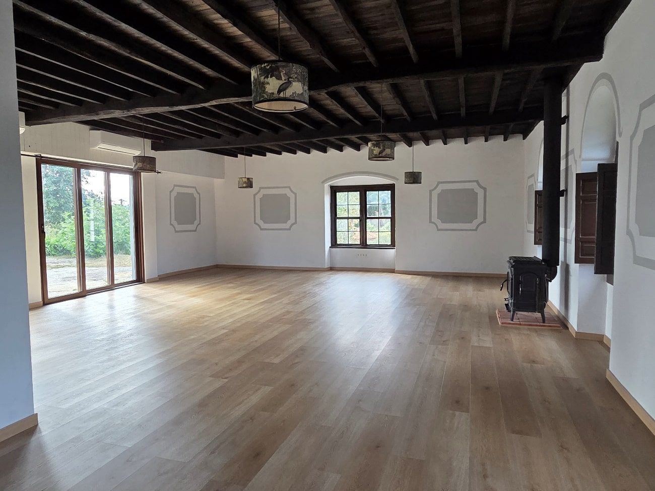 Habitación espaciosa con suelo y techo de madera, paredes blancas y grandes ventanales. Se ve una estufa de leña.