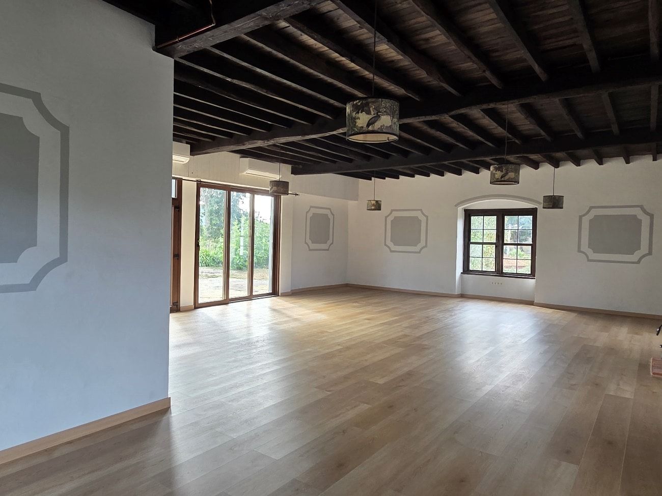Habitación espaciosa con suelo de madera, paredes blancas, vigas de madera oscura en el techo y grandes ventanales.