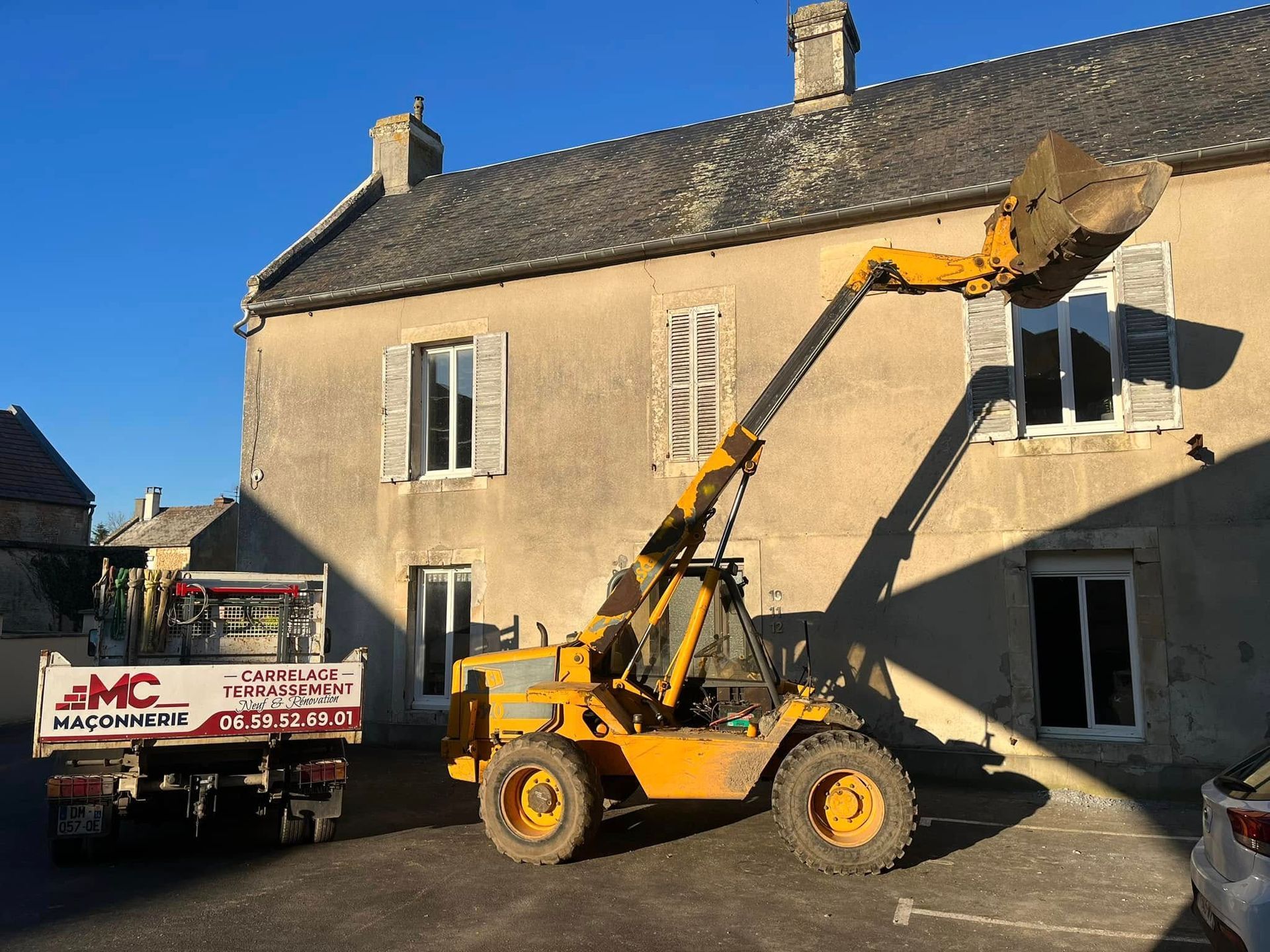 Une pelleteuse jaune travaille sur un tas de terre devant une maison blanche moderne par une journée ensoleillée.