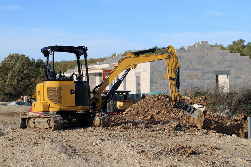 Une mini-pelle jaune creuse la terre sur un chantier de construction, devant une structure en béton partiellement construite.