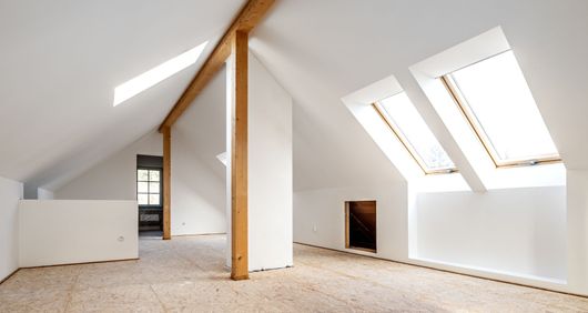 Une pièce mansardée avec un plafond blanc incliné, des poutres de soutien en bois et plusieurs puits de lumière au-dessus d'un sol clair.
