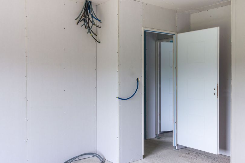 Une pièce intérieure en construction, avec des murs en plaques de plâtre blanches, des câbles apparents et une porte blanche ouverte.