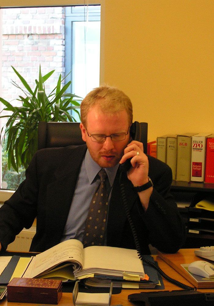 Ein Mann in Anzug und Krawatte sitzt an einem Schreibtisch und telefoniert