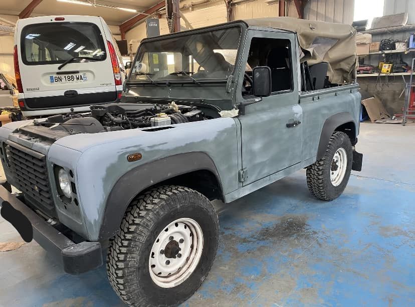 Land Rover Defender vintage gris dans un garage, vu de trois quarts avant gauche, avec une bâche souple et des pneus tout-terrain.