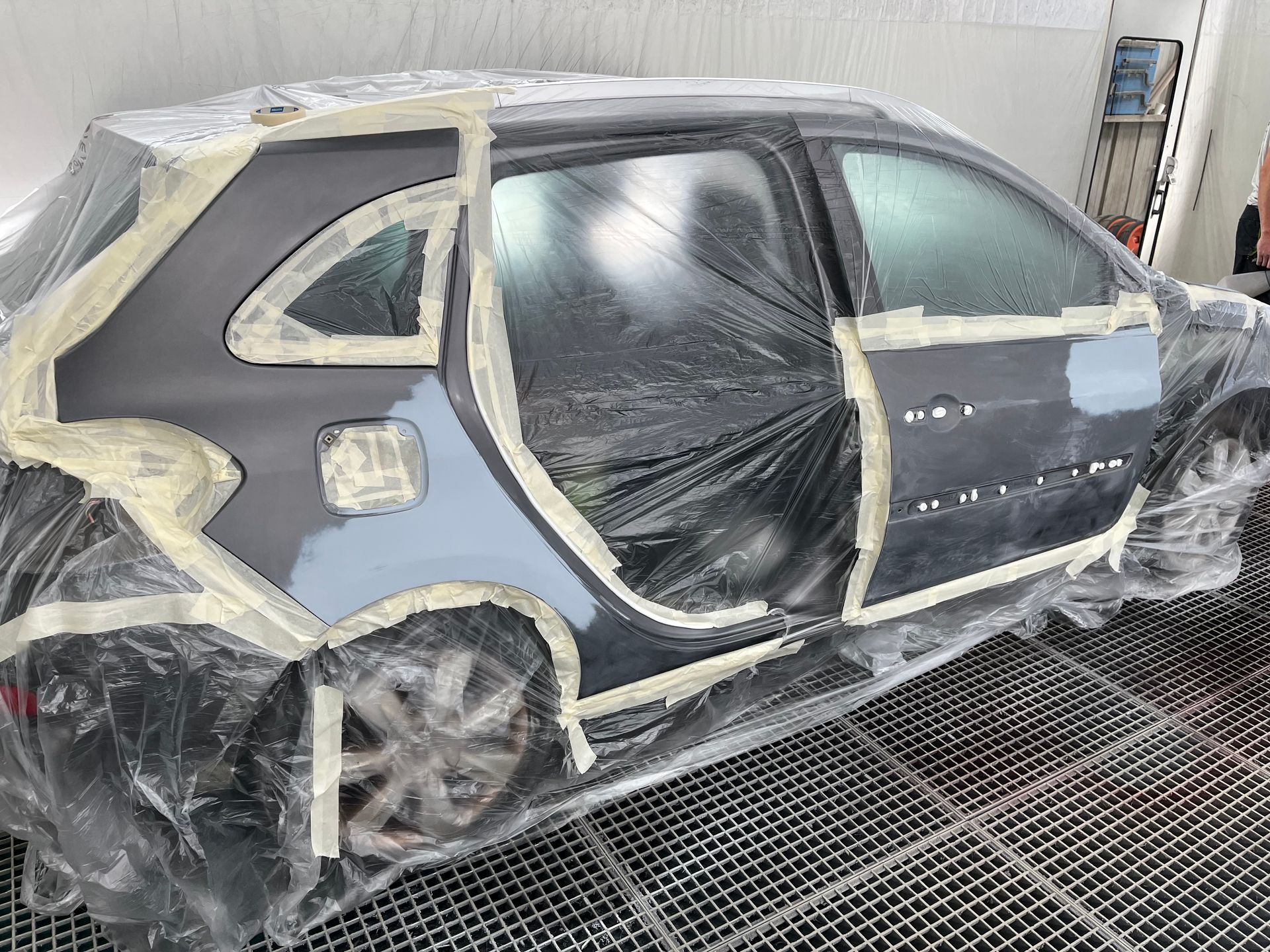 Voiture masquée avec du plastique et du ruban adhésif dans une cabine de peinture, partiellement poncée et apprêtée en vue des travaux de carrosserie.
