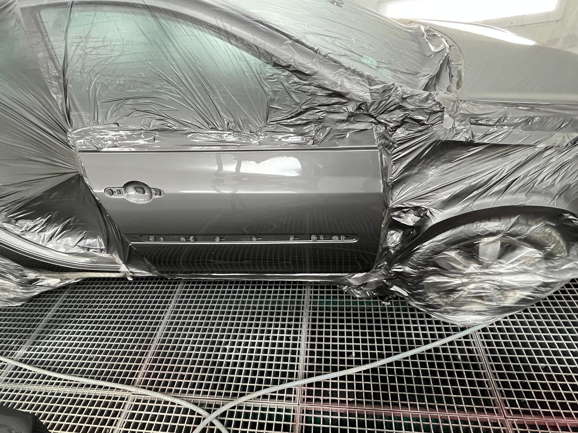 Voiture grise recouverte de plastique de masquage dans une cabine de peinture, avec des grilles de sol en dessous.